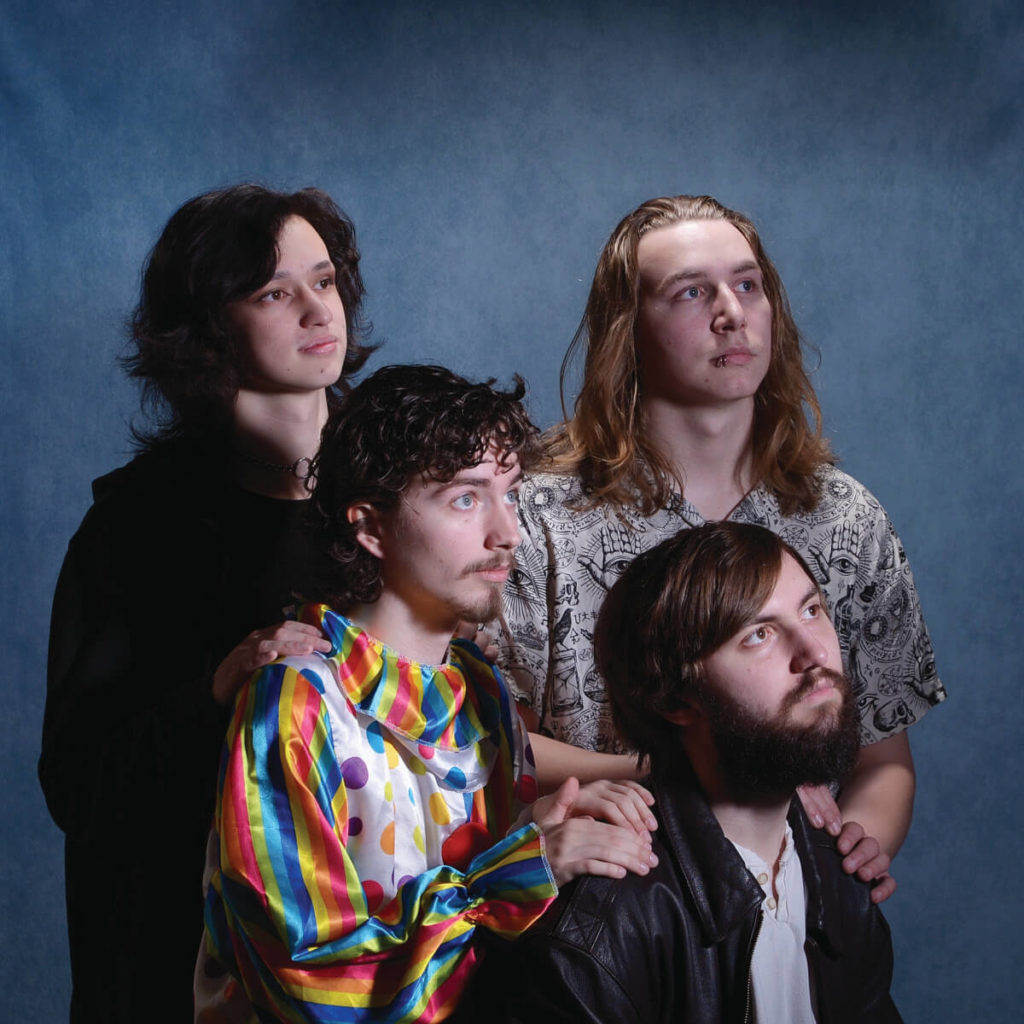 Image of four guys staring off in the distance and one is wearing a clown suite.